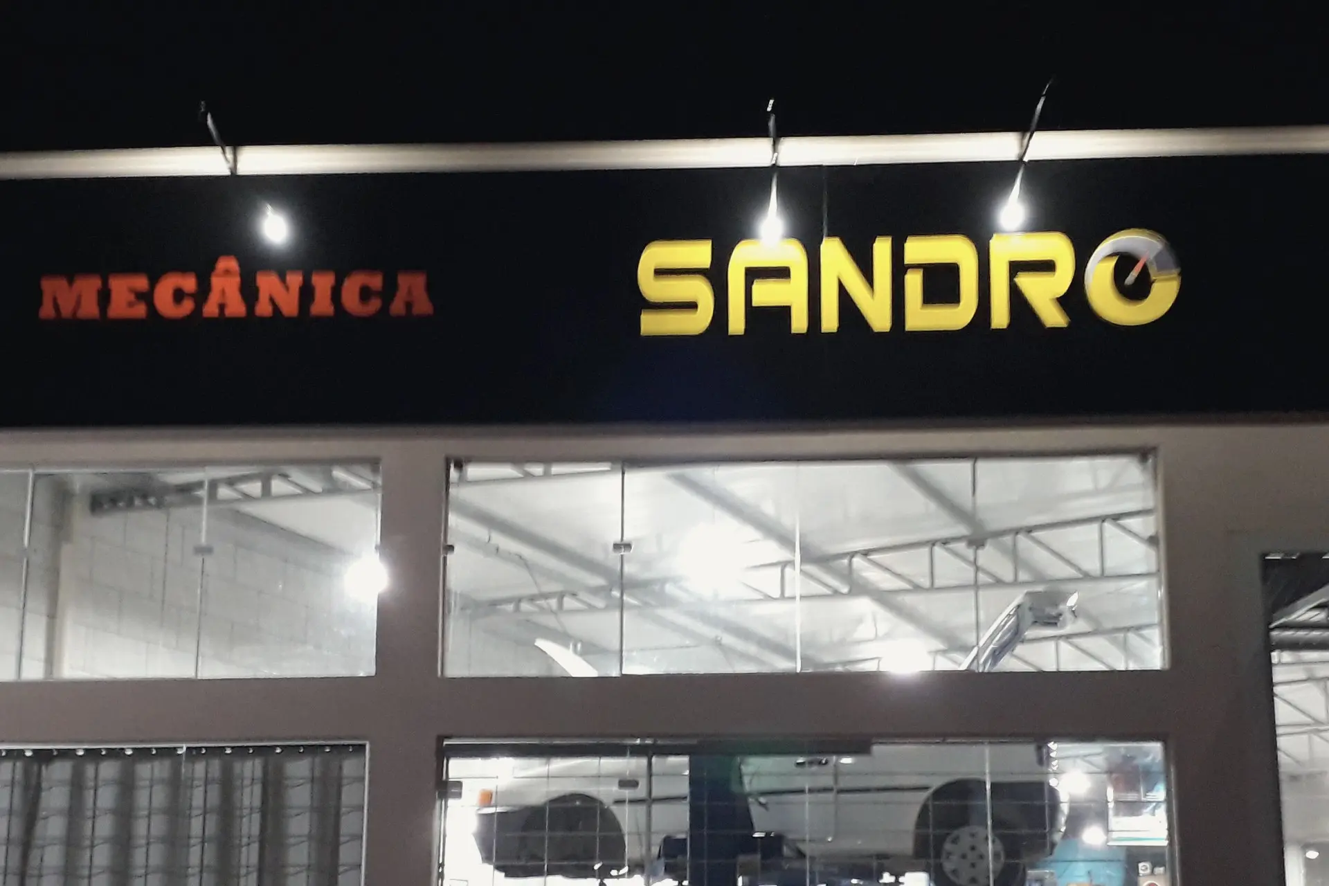 Fachada da oficina à noite, com o letreiro Mecânica Sandro aceso e, pelas vitrines, o salão com um carro no elevador